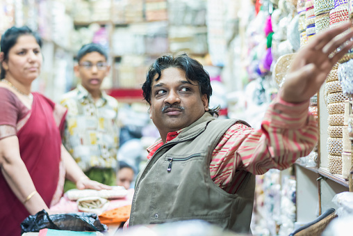 New Delhi, India - March 11, 2014: Indian shop owner sitting in  his small textile, necklaces and chain street store serving his customers, a indian woman and her son, grabbing the products out of the packed shelf.