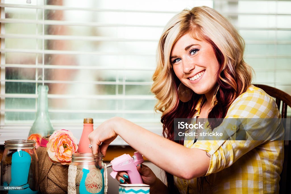 Smiling Young Woman Uses Hot-Glue Gun for Craft Project A royalty free DSLR photo of a smiling young woman as she operates a hot-glue gun to affix fabric to a mason jar painted with acrylic paint. She has blonde hair, a yellow top, and a charismatic smile. Art And Craft Stock Photo