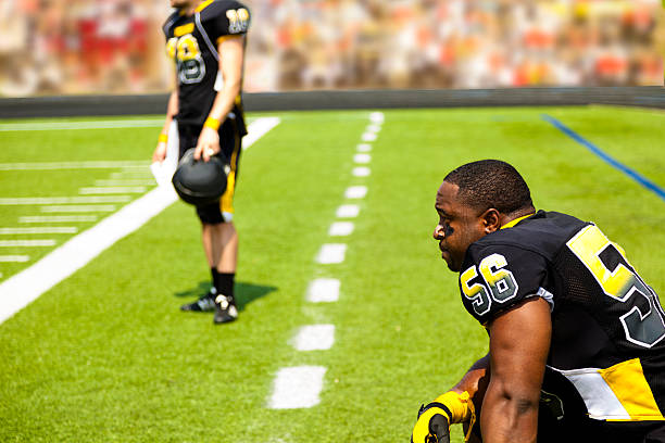Multi-ethnic group of football players wearing black and yellow uniforms waiting on the sidelines of the football field.  They wait for their turn to go into the football game. Green field with yard line markers. Fans in the background. 