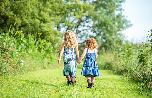 Rear view of two young girls (sisters) walking down a grass path on the farm holding hands. The older sister is on the left with curly blonde hair, a blue paisley dress, and brown cowboy boots. She is holding her younger sister's hand as they go for a walk. The younger sister has curly red hair, a blue denim dress, and brown cowboy boots. Taken ona  summer morning on the farm.