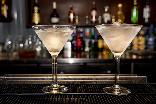 Close up photo of 2 glass dry martini on the bar. Both are ready to drink. A collection of alcohol bottle is on the photo background.