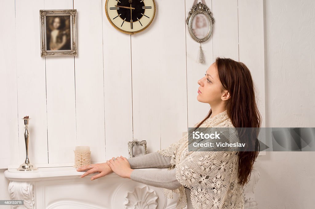 Nostalgic woman looking at an old family portrait Nostalgic young woman in a stylish shawl standing with her hands resting on a marble mantelpiece looking at an old family portrait Fireplace Stock Photo