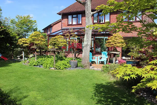 Photo showing an expanse of green lawn in the back garden of a detached red-brick house, which has been landscaped to incorporate a decking area, herbaceous orders and flower beds, trees (including Japanese maples / acer palmatum trees) and seating areas.