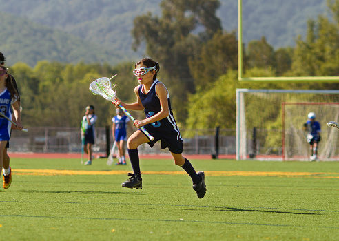 A young girl runs on a lacrosse field.  She is carrying her lacrosse stick.  The photo captures her in mid air.  