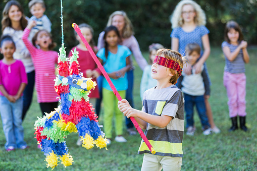 A multi-racial group of children at a birthday party, or cinco de mayo celebration, at the park.  In the foreground is a 6 year old boy, blindfolded, trying to break open a colorful pinata with a red stick while his friends watch.