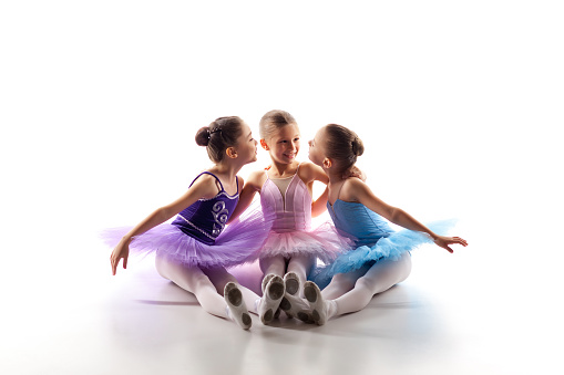 Three little ballet girls sitting  in multicolored tutu and pointe shoes together on white background