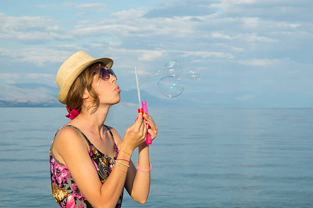 Young girl blowing soap bubbles at the beach by the sea wearing straw hat and sunglasses