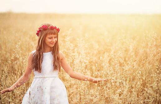 Little girl aged 9 wearing white dress and wreath walking in oat grain field. 