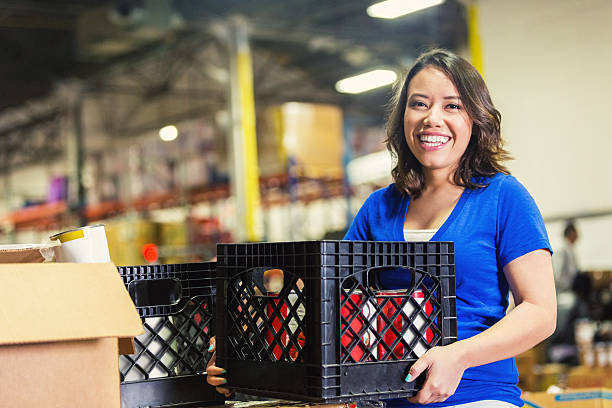 Young Asian American volunteer working in food bank warehouse Young adult Asian and Hispanic American woman is smiling and looking at camera. She is holding a crate of donated grocery and food items. She's volunteering in a large food bank warehouse. Woman is sorting donated food to distribute to community. grocery shelf cans stock pictures, royalty-free photos & images
