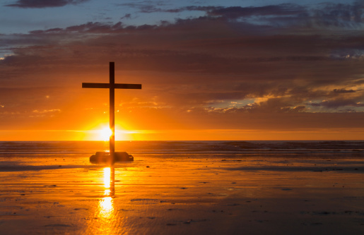 Cross on a beach with the sun setting down behind it.