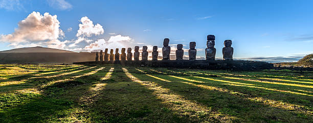 Panorama of Ahu Tongariki at sunrise, Easter Island, Chile. Panoramic view of the 15 Moai statues at Ahu Tongariki at sunrise, Easter Island (Rapa Nui), Chile. The backlit statues, against a bright blue sky with white clouds, cast long shadows upon the green grassy field in the foreground. easter island stock pictures, royalty-free photos & images
