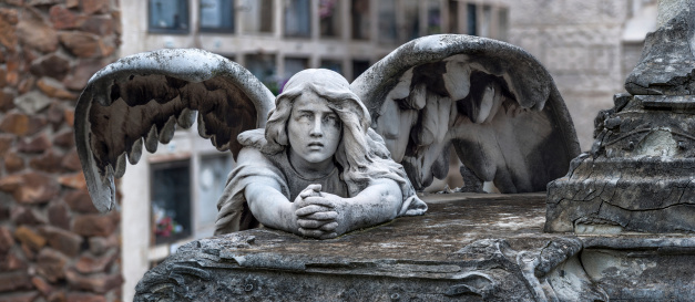 Panoramic close up of a beautifully crafted white marble sculpture at the Cemetery of Montjuich, Barcelona, Spain. The sculpture, created in the early 1900s, depicts sorrow, or grief, in the form of a winged angel kneeled down over a tomb with it's hands clasped in prayer. In the foreground, the cracked, weathered stone-surface of the tomb evidences it's age. In the blurred out background appears a wall containing above ground burial vaults.  