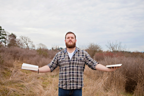 A young man holds two open Bibles in his hands. Unsure which to follow? Comparing the differences? On a search.