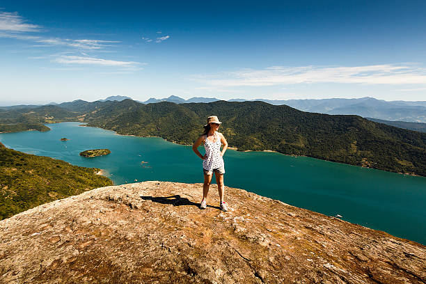 Young Woman on Top of High Mountain on Sunny Morning A DSRL photo taken at the top of a mountain beside the calm sea in Mamangua, Paraty, Rio de Janeiro, Brazil. At the very top there is a young caucasian woman wearing hat and sunglasses contemplating the view. It is a sunny day with bright blue sky.  paraty brazil stock pictures, royalty-free photos & images