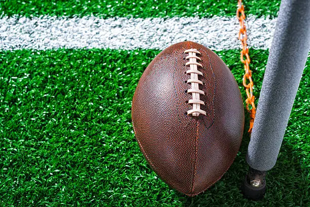 Looking down on an American Football first down, the nose of the ball just past the pole of chain marker with a yard line on the artificial turf.