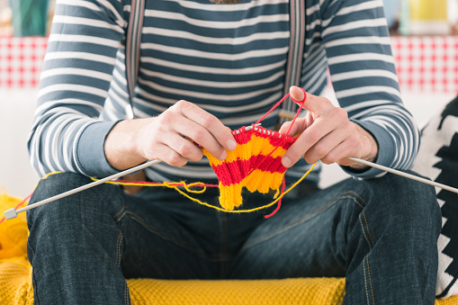 Young man wearing striped top sitting on sofa at home and knitting. Close up of hands, unrecognizable person.
