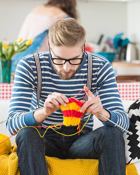Young bearded man knitting Young bearded man wearing nerd glasses and striped top sitting on sofa at home and knitting, with his girlfriend in the kitchen in the background. knitting needle photos stock pictures, royalty-free photos & images
