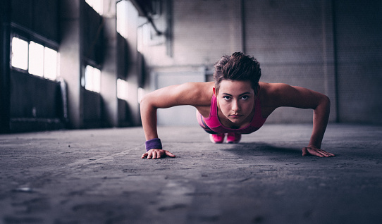 Determined teen girl looking at the camera with a focused expression while doing push ups on the grey concrete floor of a grungy industrial space
