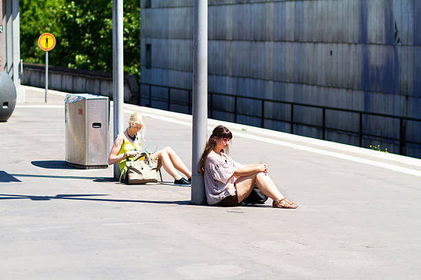 Essen, Germany - June 30, 2015: Cature of two young caucasian women waiting for train in station Essen. Hot summer shot. Girls are sitting on floor at lanterns in sunshine.