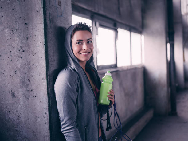 Teen girl smiling holding water bottle after a working out Pretty teen girl in a grey hoodie smiling holding a green water bottle after a working out in a grungy industrial space crossfit girl stock pictures, royalty-free photos & images