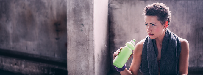 Banner style image of a pretty young woman with a serious expression with a towel over her shoulders and holding up a water bottle while taking a break ofter a gym workout