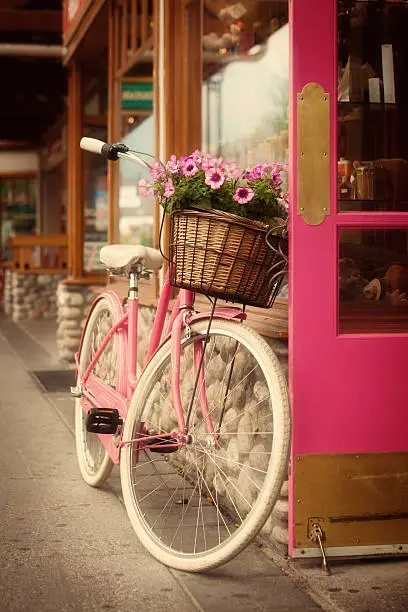 A nostalgic scene of an old fashion pink bicycle with a flower basket in the front. The bike is parked and leaning against the pink door of a retail shop along the pedestrian sidewalk. An old fashion retro scene of the past. Photographed in vertical format and processed with de-saturation and warm tone.