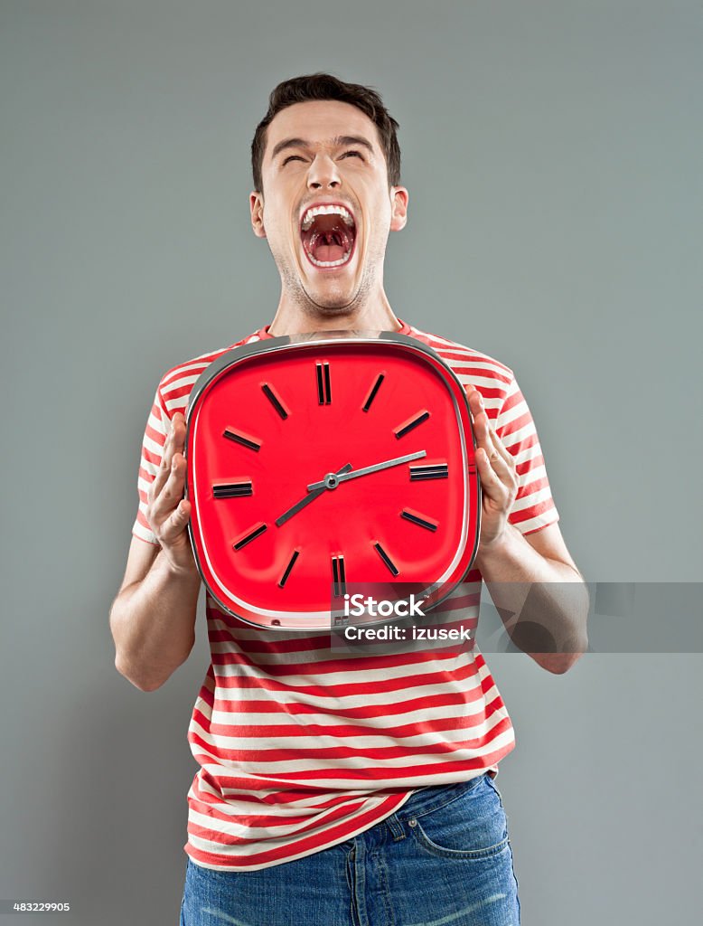 Young man with red clock Portrait of excited young man wearing striped t-shirt, holding a big red clock and screaming. Studio shot, grey background. Young Men Stock Photo