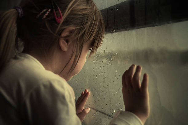 Lonely child looking through window Sad little girl looking through the rainy window. Grain added for texture. child abuse photos stock pictures, royalty-free photos & images
