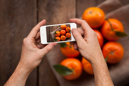 A studio shot image of a male adult caucasian hand taking a selfie of a group of mandarins, using a smart phone. 