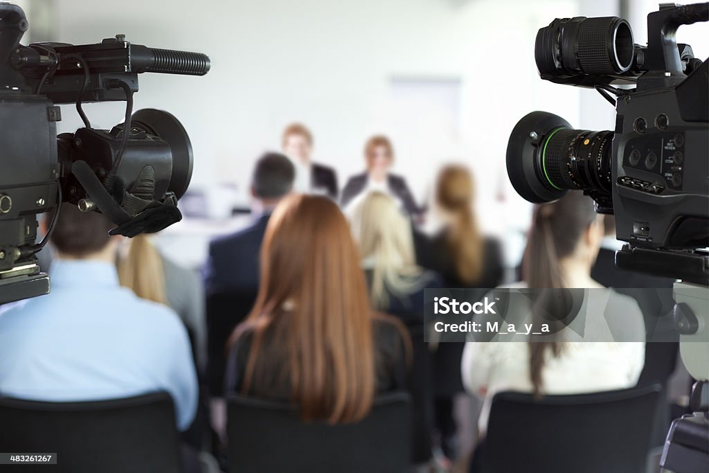 Press conference. Cameras pointed at man and women giving a presentation in background. Focus on cameras. Adult Stock Photo