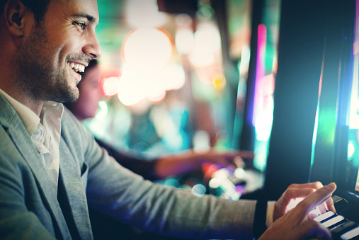 Closeup of handsome earlu 30's smiling man playing slots in a casino. He's wearing gray suit jacket and white shirt with unbuttoned collar. Having great time and making some money. There are blurry people in background also playing slots.