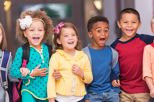 A multi-ethnic group of children standing in a row in a school hallway, excited and laughing, watching something.  They are in kindergarten or preschool, carrying bookbags.  They are 4 to 6 years old.