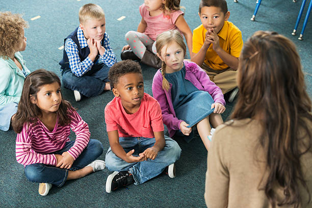 Kindergarten students listening to teacher A group of multiracial kindergarten students sitting on the floor in a classroom, looking up at the teacher who is sitting on a chair in front of them.  They have serious expressions on their faces. sitting on floor stock pictures, royalty-free photos & images