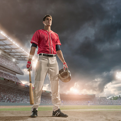 A low angle portrait image of a professional baseball player – hitter - dressed a full generic baseball kit, pads, gloves and holding baseball bat looking off camera into the distance. The batter is standing on a baseball field in a generic floodlit outdoor baseball stadium full of spectators under a dark stormy evening sky. 