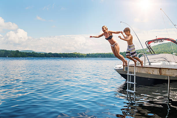 Mother and son jumping in lake from pier sunny day. Playing in water is a summer treat for families. This mother and her son are jumping in lake from pier on sunny day. This was taken in the Laurentian forest of Quebec, Canada. Water is calm, sun is shining, boat is attached to the pier. Full length horizontal shot with copy space. boat on lake stock pictures, royalty-free photos & images