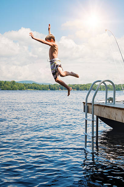 Playing in water is a summer treat for children. This little boy is jumping in lake from pier on sunny day. This was taken in the Laurentian forest of Quebec, Canada. Water is calm, sun is shining. Full length vertical shot with lens flare and copy space.
