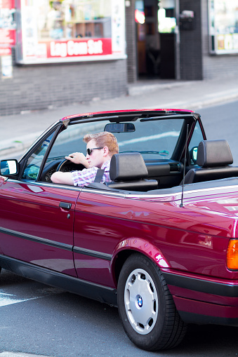 Essen, Germany - July 1, 2015: A young caucasian adult is sitting in bordeaux colored BMW convertible car and is wearing sunglasses. Summertime shot. Man is checking street and traffic from left side before turning to right side.