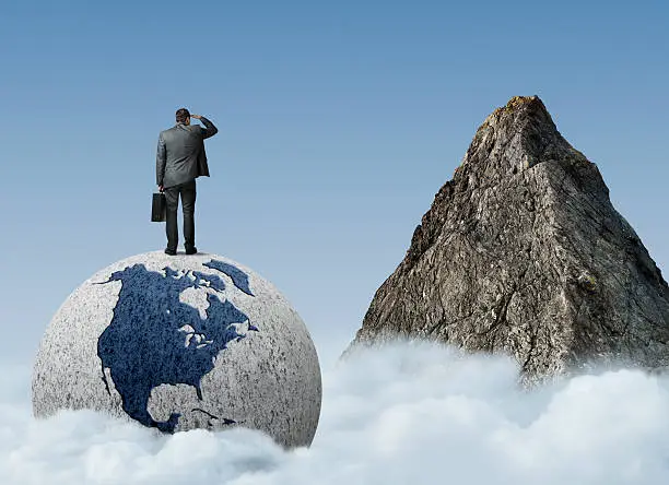 A businessman standing on top of a globe staring up at a mountaintop.
