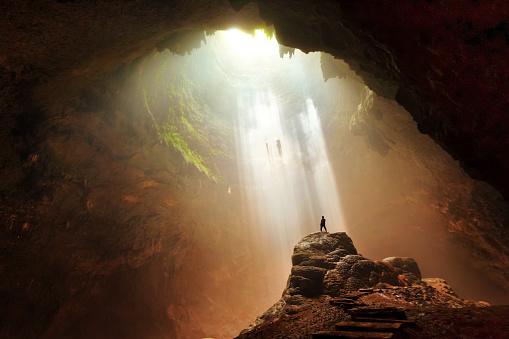 A man standing on stone undergound cave. Ray of Light of heaven was taken in Jomblang Cave,  a vertical cave with dense ancient forest below. A 300 meter alley with its beautiful natural ornament a place where can see light from heaven.