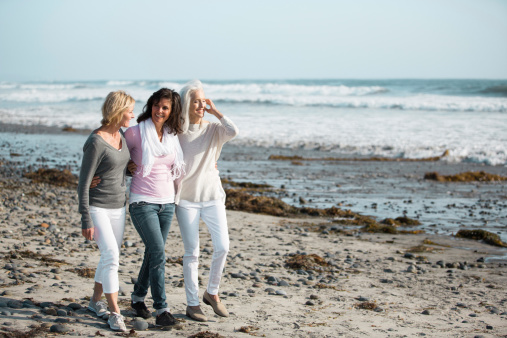 Three smiling mature women embracing one another on a rocky, seaweed covered sandy beach. The ocean and clear blue skies are behind them. Women have been friends for years and share a deep connection. 