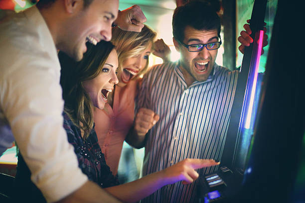 Group of young adults having fun in casino. Group of young adults in late 20's playing slot poker and fruits and wining.Wearing casual clothes and having fun on weekend night. There are many slot machines out of focus in background. arcade stock pictures, royalty-free photos & images