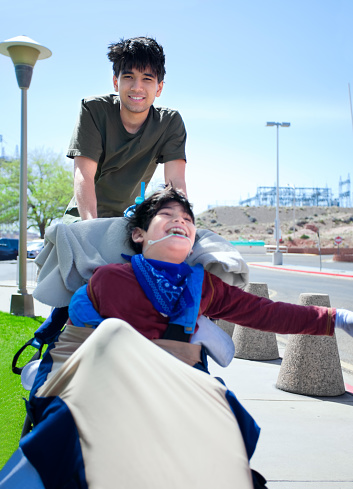 Teen boy pushing happy disabled biracial little  brother in wheelchair, smiling and laughing, outdoors. cerebral palsy