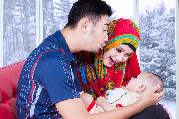 Portrait of two young muslim parents playing with their baby on sofa at home, shot with winter background on the window