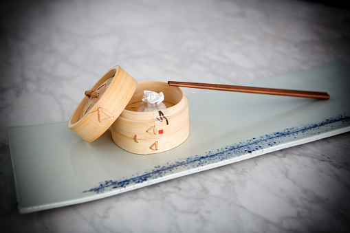 Dim sum appetizer consisting of a single pork bun in a traditional bamboo steamer basket and one set of chop sticks.
