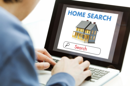 An unidentifiable man searching online for a new home, using a laptop computer internet web real estate listing search engine tool. His hands tap the keyboard as a typical, generic house residential structure and the text “Home Search” appear on the monitor screen. People shop using technology for e-commerce convenience.