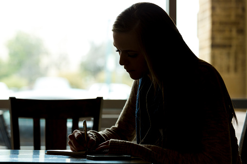 Silhouette of young woman writing alone in a coffee shop.