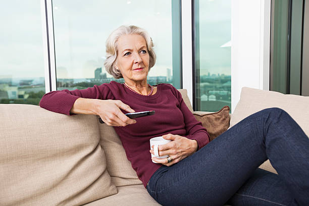 istock Senior woman using TV remote control on sofa at home 483390207