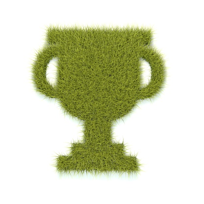 The shape of a trophy is made out of lush green grass. The shape is on a plain white background with some shadows.