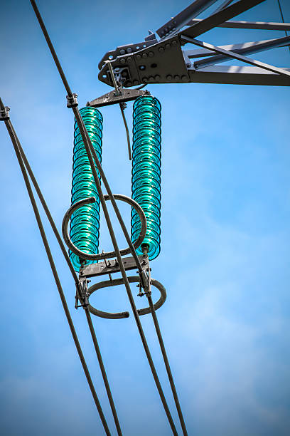 Vertical composition photography shot without people, of close-up of glass insulator circles from high voltage metallic electricity pylon with power line cable. This image was taken against blue sky, with a small vignetting effect.