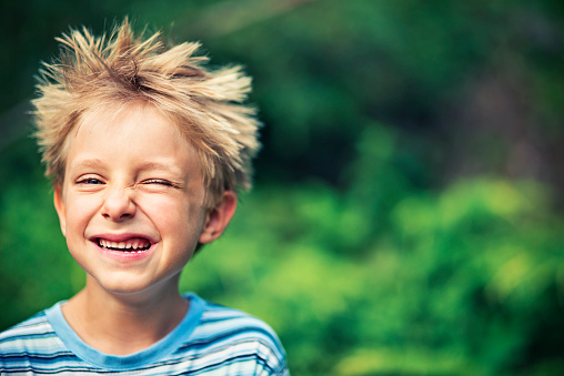 Portrait of a funny little boy wiking and laughing. The boy is aged 5 and is standing outdoors smiling at the camera trying to wink.